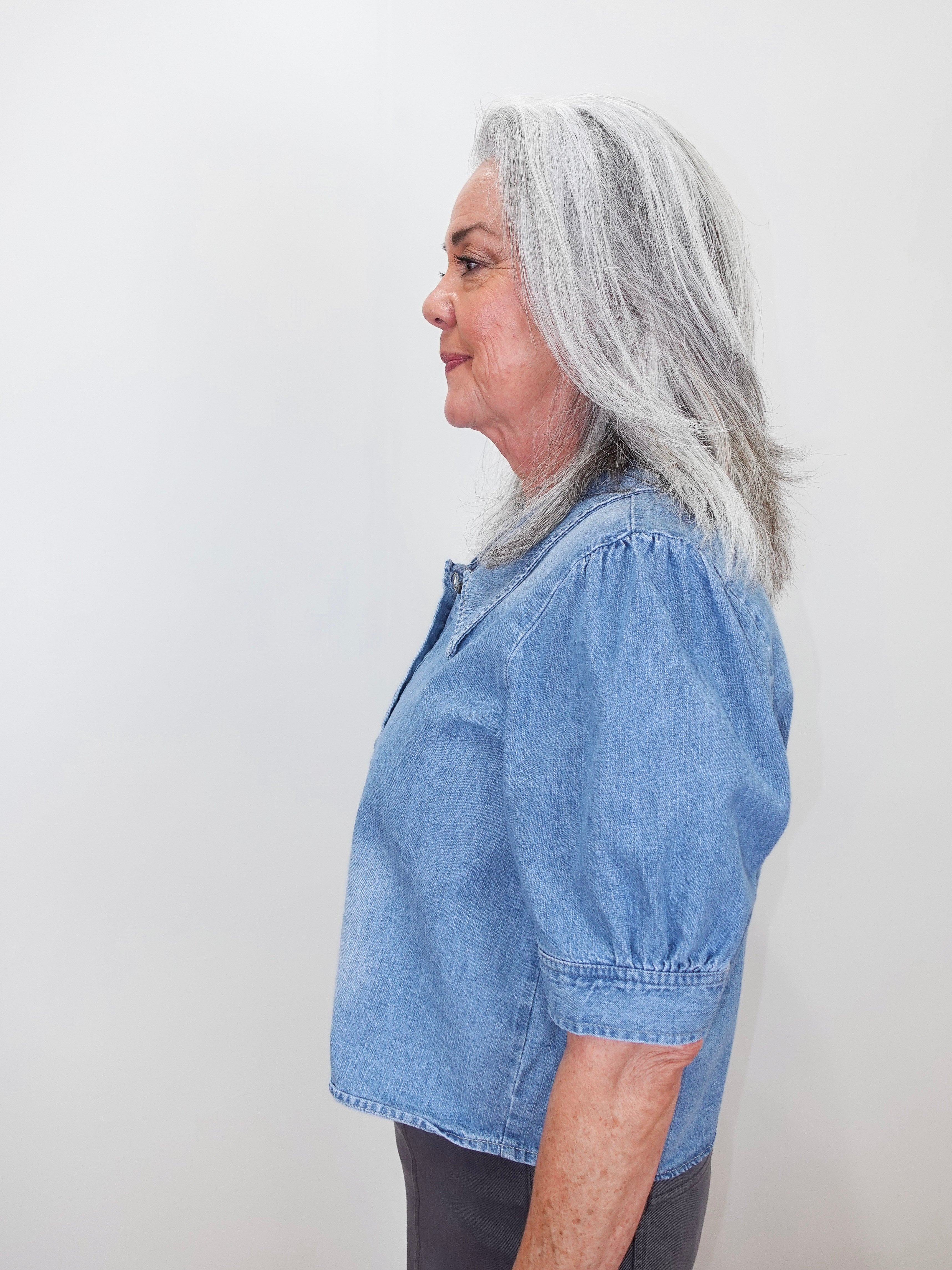 A woman with shoulder-length gray hair is shown in profile wearing a light blue cropped short-sleeve blouse with puff sleeves and dark gray pants, standing against a plain off-white wall.