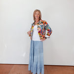Woman wearing a floral jacket, white top, and blue skirt against a white wall.