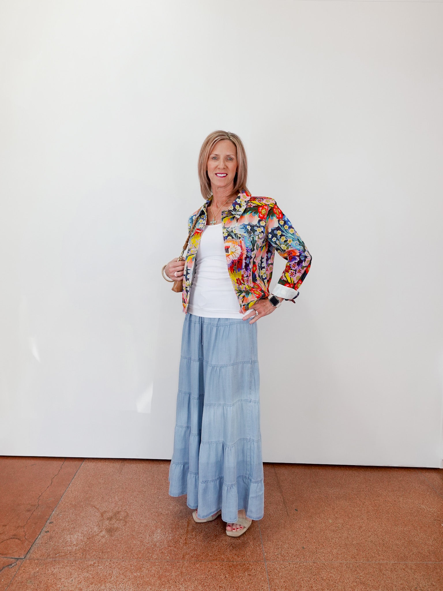 Woman wearing a floral jacket, white top, and blue skirt against a white wall.