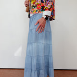 Person wearing a colorful floral jacket and blue tiered skirt against a white wall.