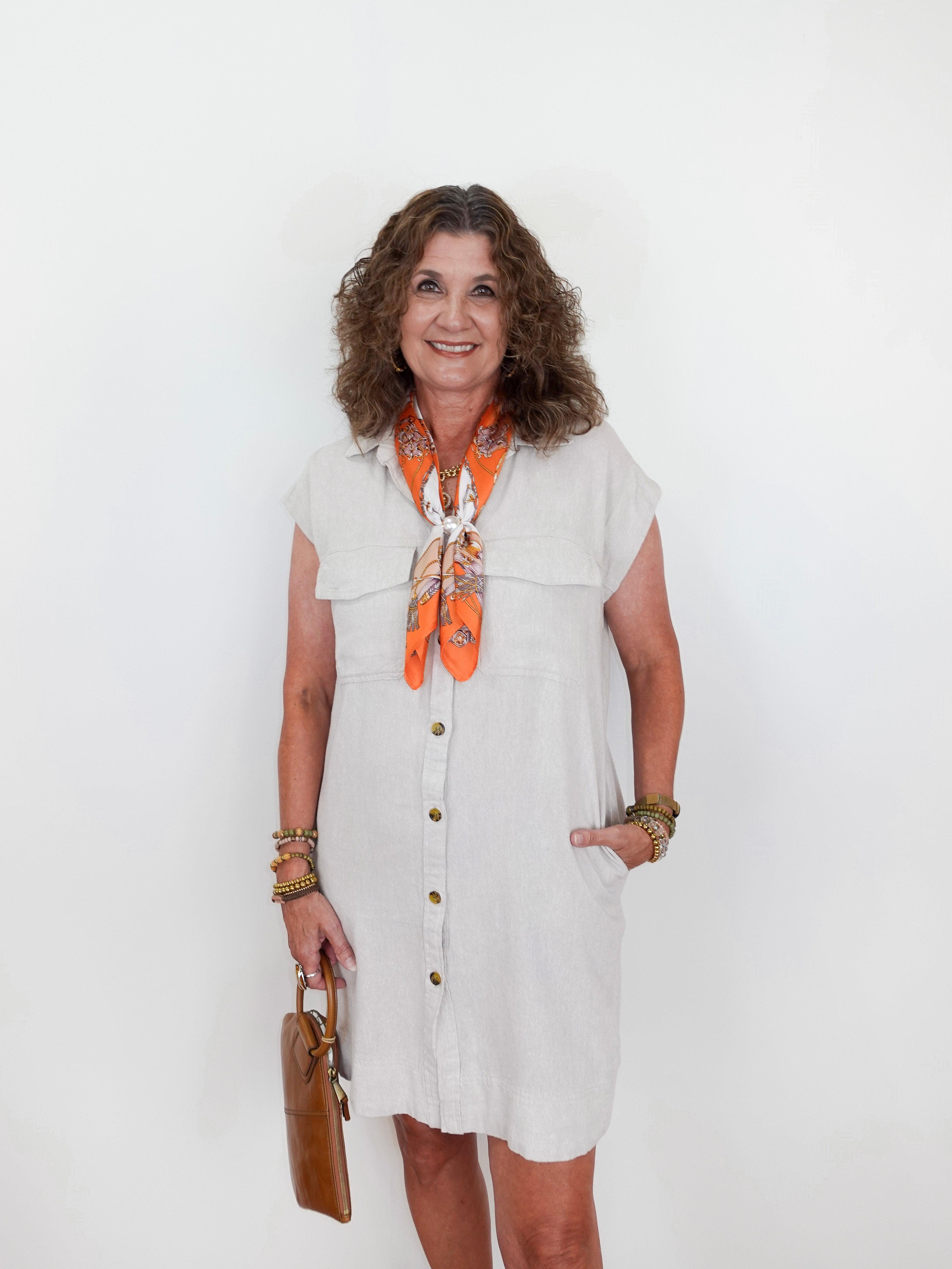 Woman wearing a white dress with an orange scarf, holding a brown bag against a white background