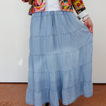 Person wearing a colorful floral jacket, white shirt, and blue tiered skirt on a plain background