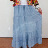 Person wearing a colorful floral jacket, white shirt, and blue tiered skirt on a plain background