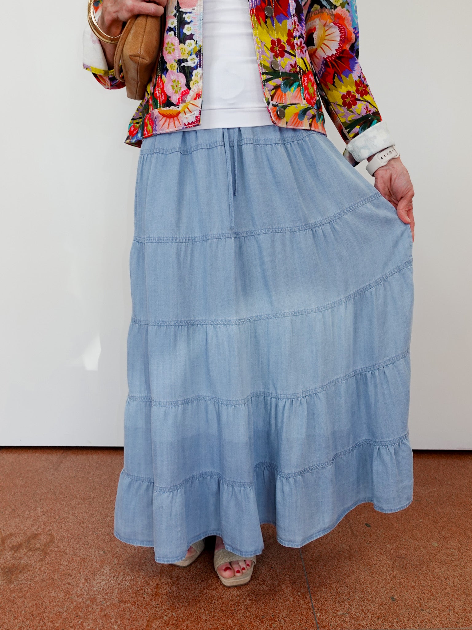 Person wearing a colorful floral jacket, white shirt, and blue tiered skirt on a plain background