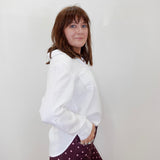 A woman with brown hair is shown from the waist up, wearing a white long-sleeve button-up shirt and dark burgundy pants with a light pink polka-dot pattern, standing against a plain off-white wall.