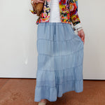 Person wearing a colorful jacket, white top, blue tiered skirt, and beige shoes on a plain background