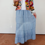 Person wearing a colorful jacket, white top, blue tiered skirt, and beige shoes on a plain background