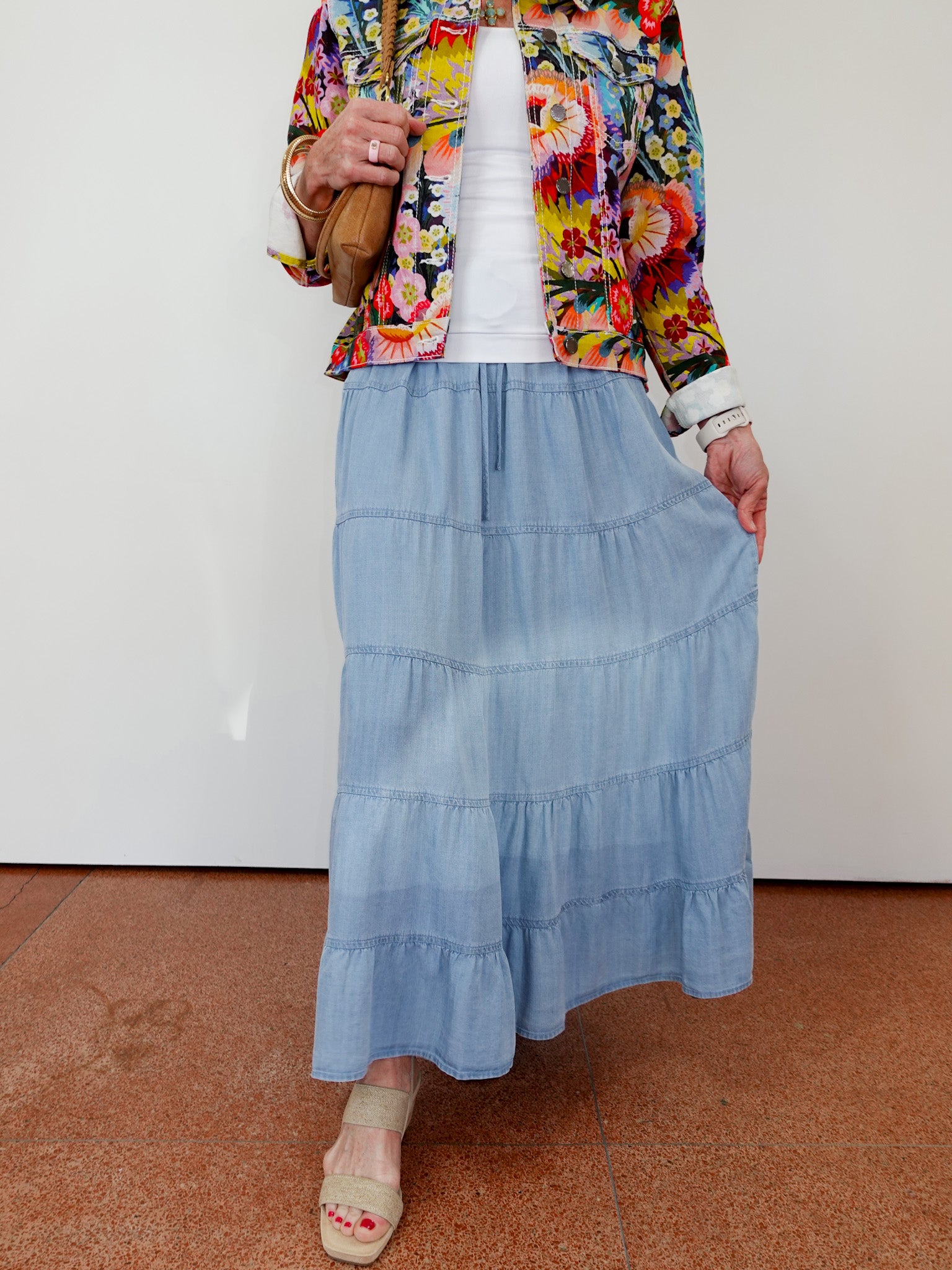 Person wearing a colorful jacket, white top, blue tiered skirt, and beige shoes on a plain background