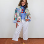 Woman wearing a colorful patchwork jacket and white pants against a white wall.