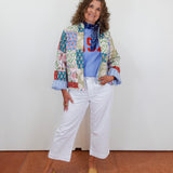 Woman wearing a colorful patchwork jacket and white pants against a white wall.