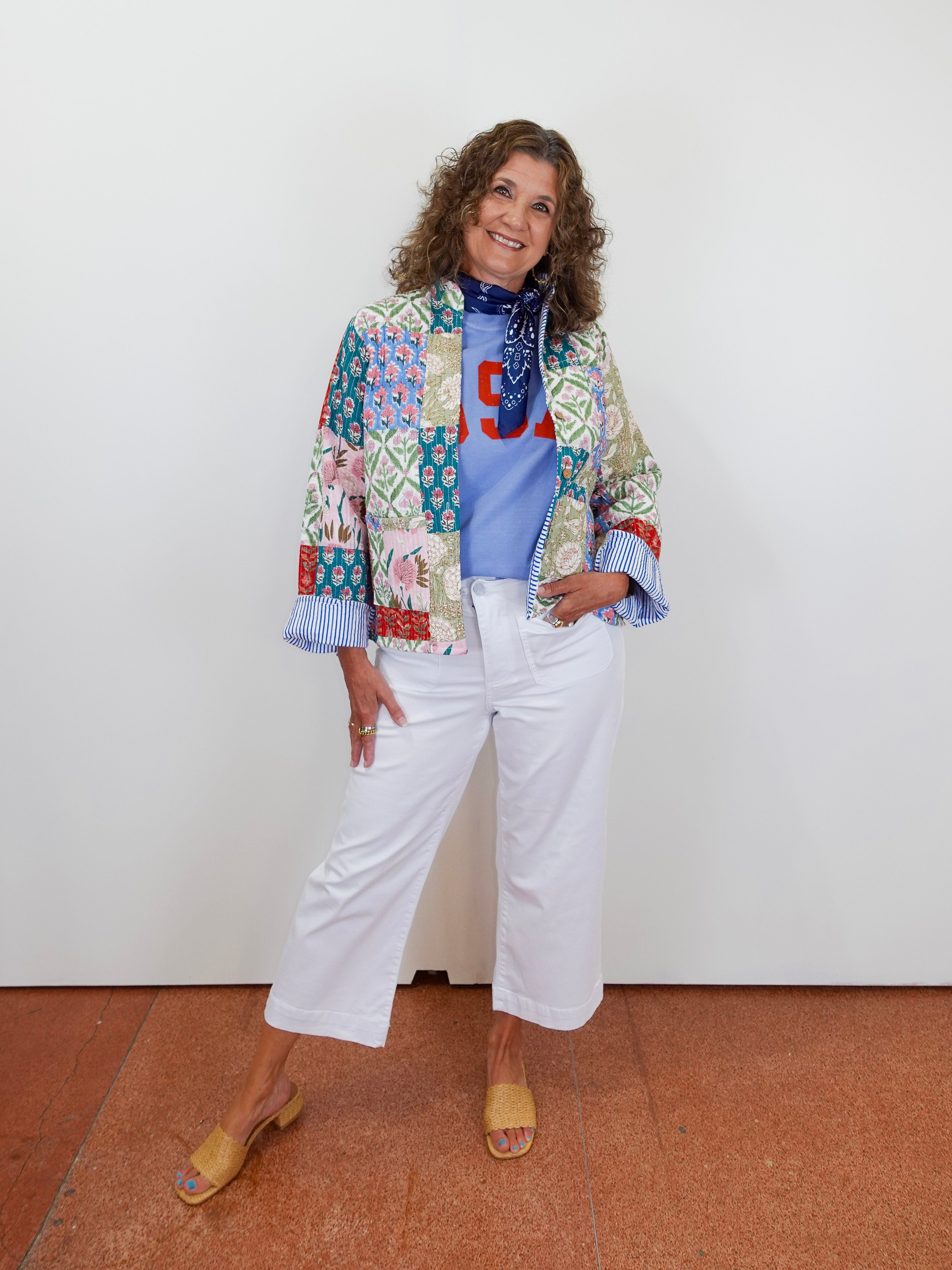 Woman wearing a colorful patchwork jacket and white pants against a white wall.