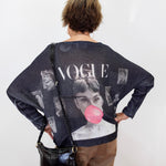 A woman viewed from behind wearing a dark navy blue knit sweater with a large graphic print featuring Audrey Hepburn, the word 'VOGUE', and a pink bubblegum bubble motif. She is also wearing brown pants and carrying a black shoulder bag against a plain white wall.