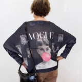 A woman viewed from behind wearing a dark navy blue knit sweater with a large graphic print featuring Audrey Hepburn, the word 'VOGUE', and a pink bubblegum bubble motif. She is also wearing brown pants and carrying a black shoulder bag against a plain white wall.