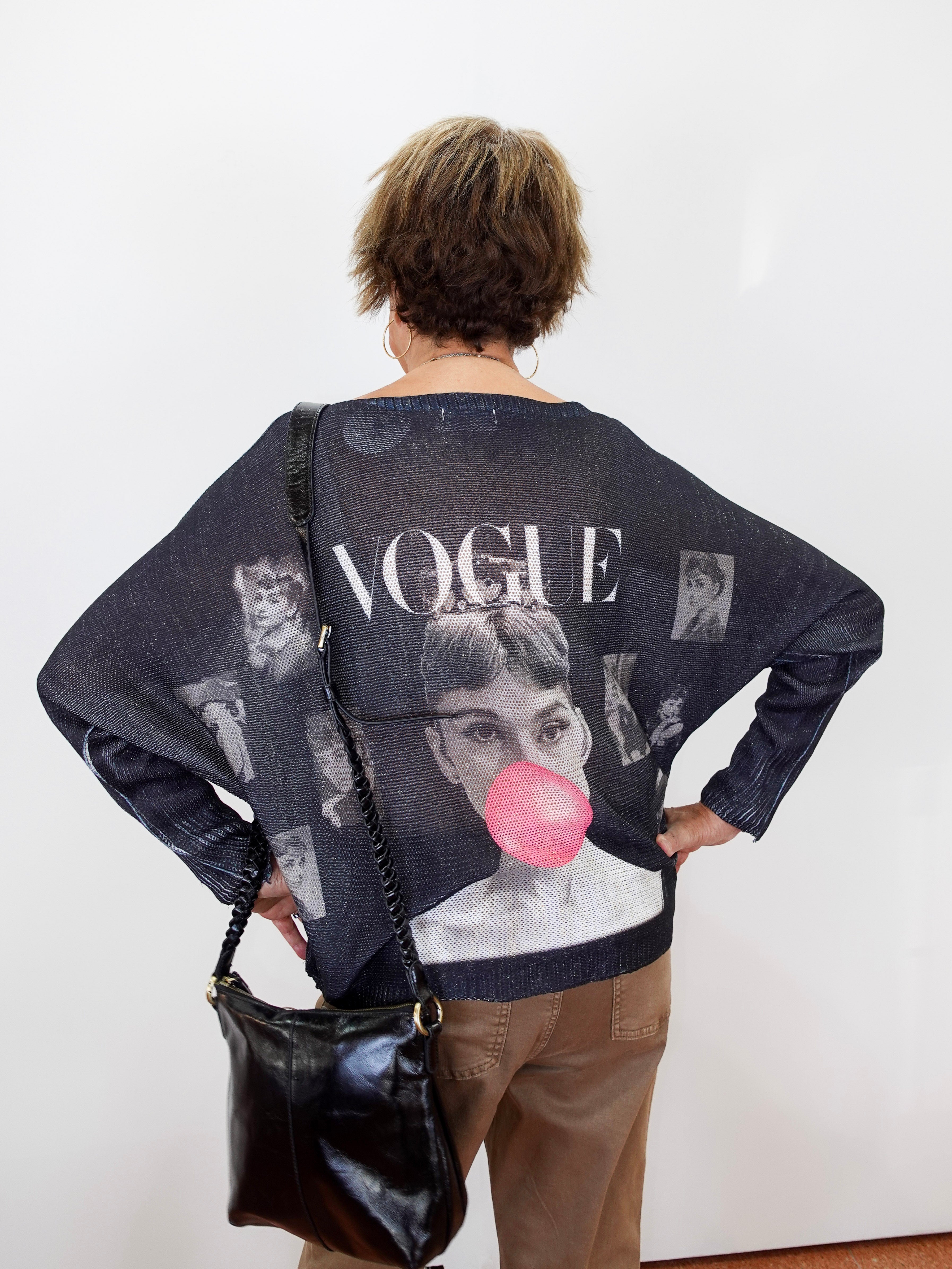 A woman viewed from behind wearing a dark navy blue knit sweater with a large graphic print featuring Audrey Hepburn, the word 'VOGUE', and a pink bubblegum bubble motif. She is also wearing brown pants and carrying a black shoulder bag against a plain white wall.