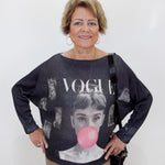 An older woman stands against a plain white wall wearing a dark gray oversized knit sweater with a large graphic print featuring the word 'VOGUE' and a pink bubblegum bubble motif. She is also wearing tan pants and carrying a black shoulder bag.