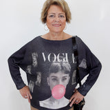 An older woman stands against a plain white wall wearing a dark gray oversized knit sweater with a large graphic print featuring the word 'VOGUE' and a pink bubblegum bubble motif. She is also wearing tan pants and carrying a black shoulder bag.