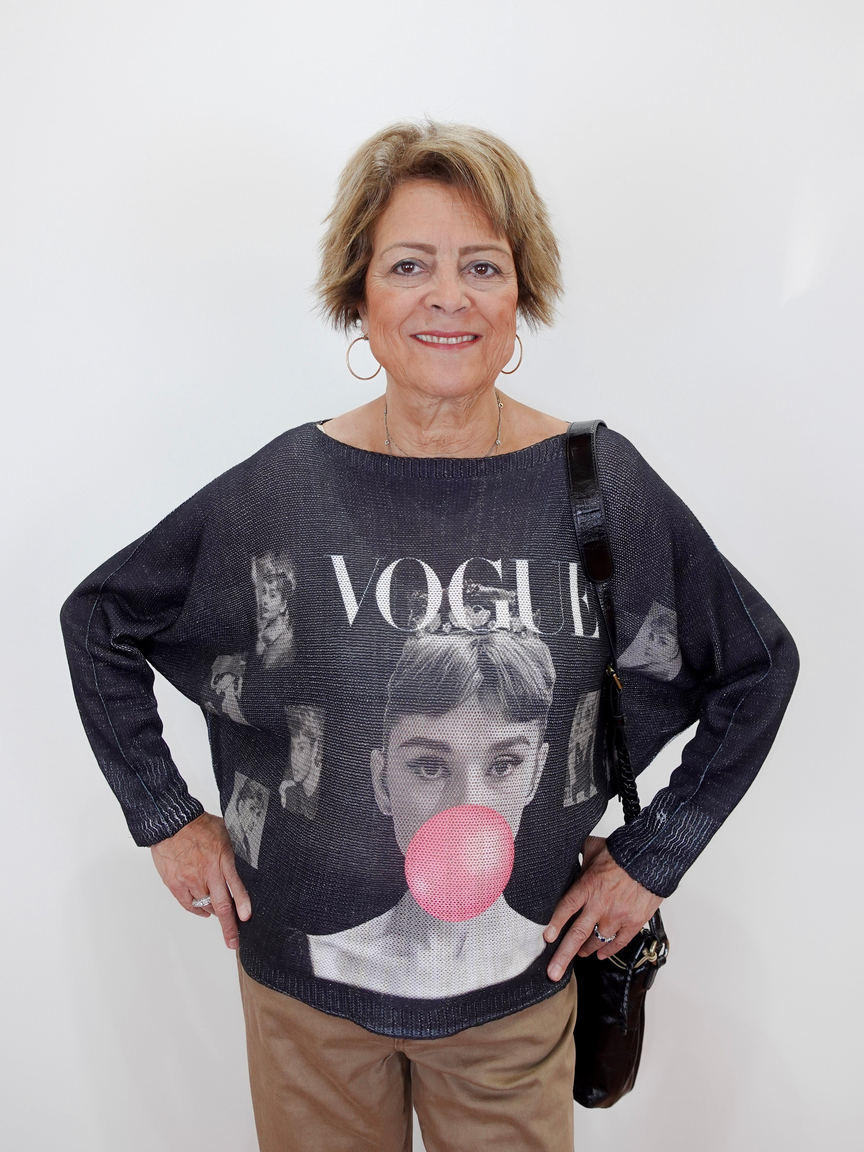 An older woman stands against a plain white wall wearing a dark gray oversized knit sweater with a large graphic print featuring the word 'VOGUE' and a pink bubblegum bubble motif. She is also wearing tan pants and carrying a black shoulder bag.