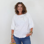 Woman wearing a white blouse and blue jeans holding a decorative bag against a plain background