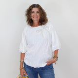 Woman wearing a white blouse and blue jeans holding a decorative bag against a plain background