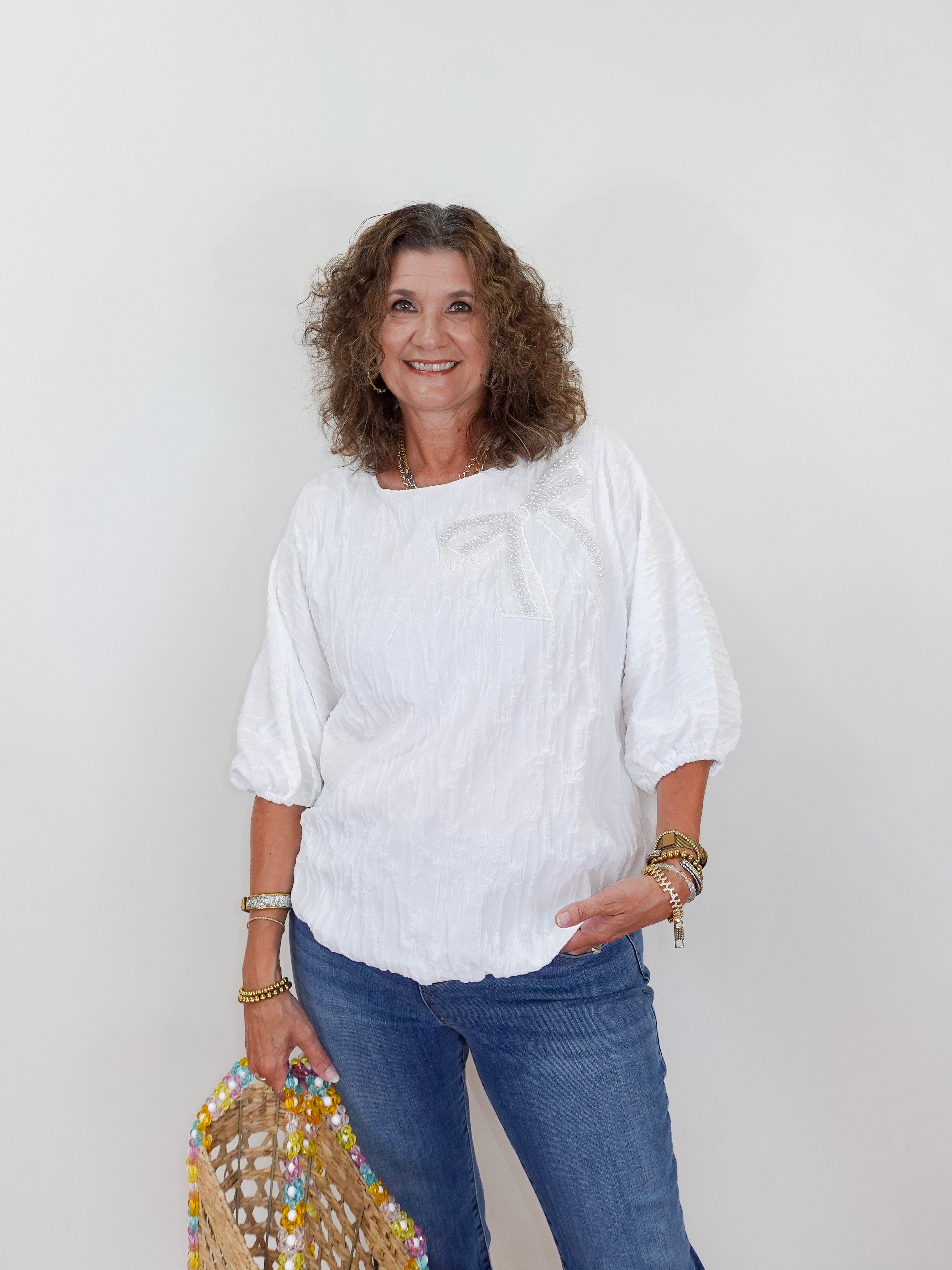 Woman wearing a white blouse and blue jeans holding a decorative bag against a plain background