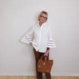 Woman wearing a white blouse with bell sleeves, brown pants, and holding a brown handbag against a plain background.