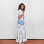 Woman in a floral dress holding a blue clutch against a white wall.