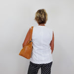 Person wearing a white sleeveless top and black patterned pants, holding a brown bag against a plain background