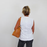 Person wearing a white sleeveless top and black patterned pants, holding a brown bag against a plain background