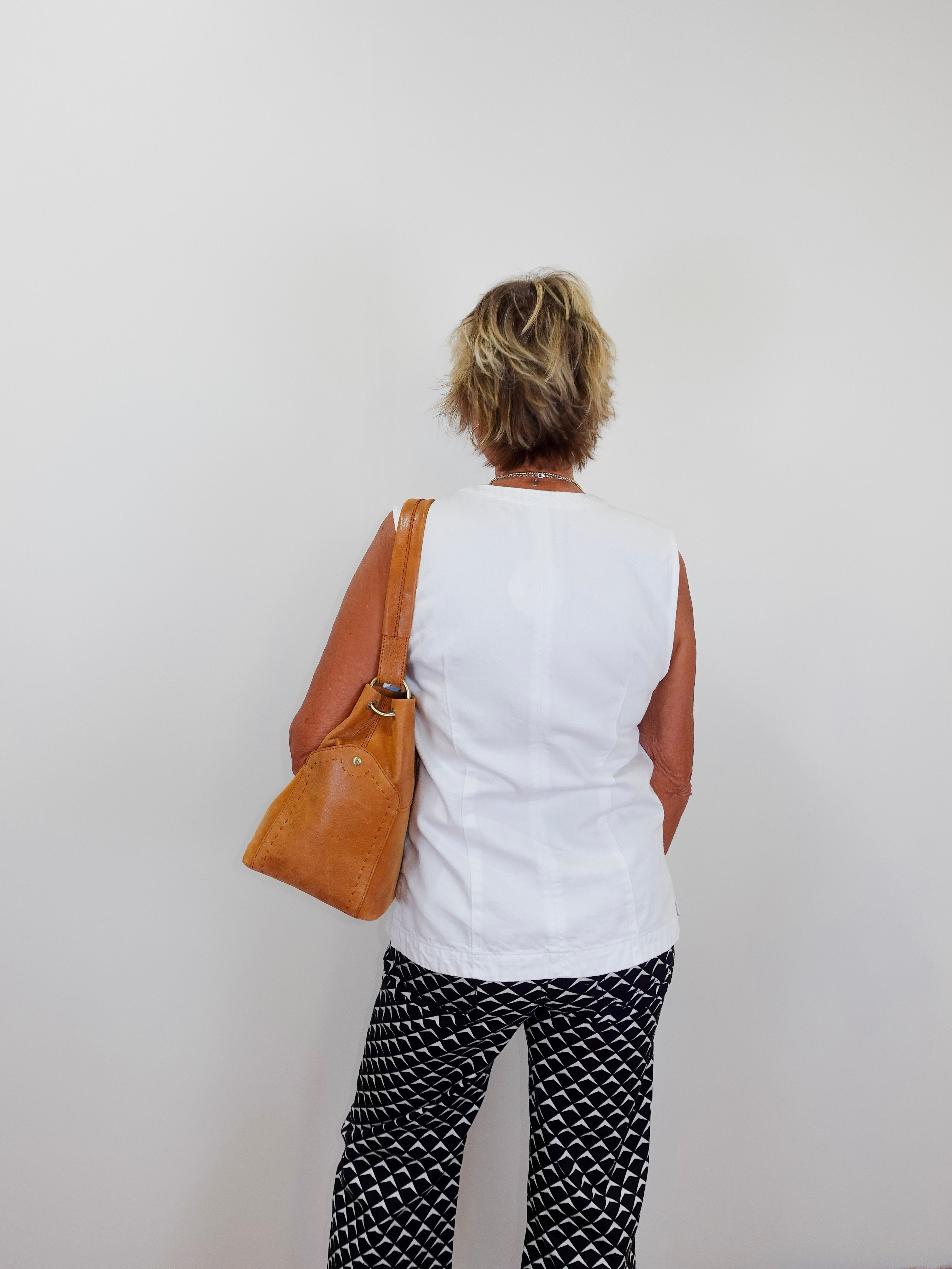 Person wearing a white sleeveless top and black patterned pants, holding a brown bag against a plain background