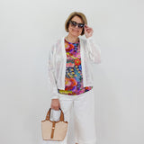 Woman wearing a colorful top, white pants, and a white cardigan against a plain background