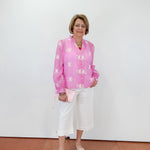 Woman wearing a pink floral blouse and white pants against a white wall.