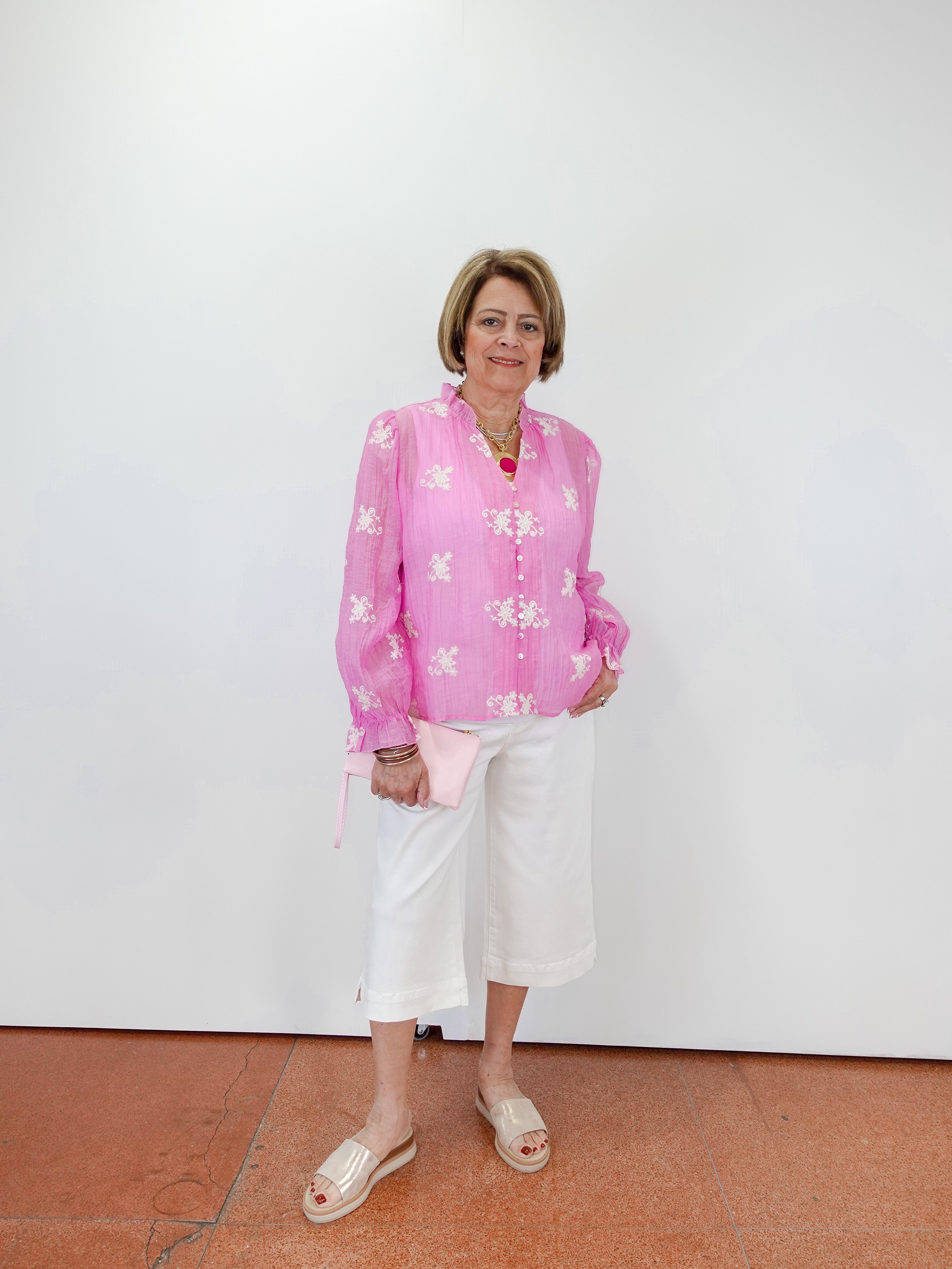 Woman wearing a pink floral blouse and white pants against a white wall.