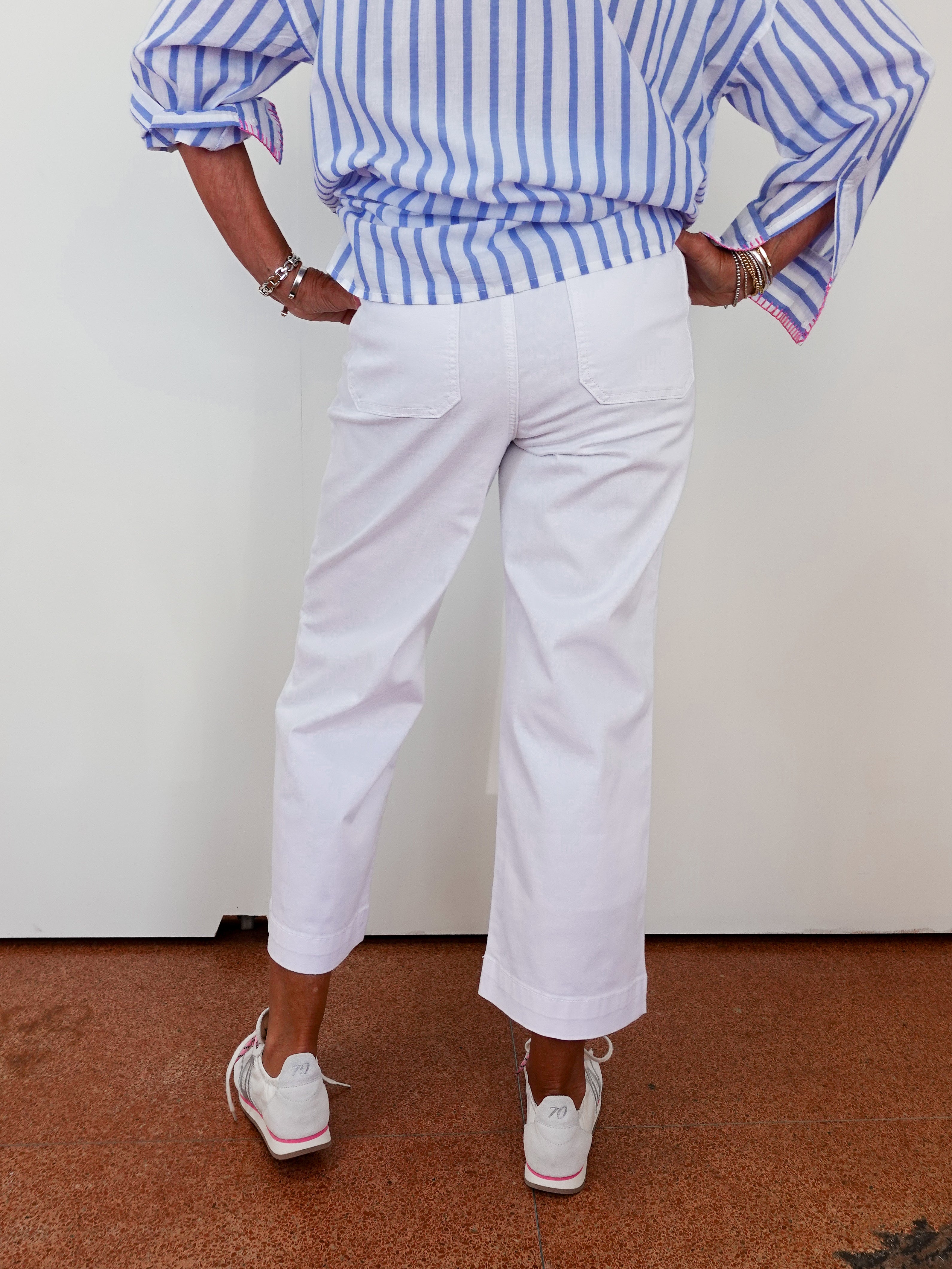 Person wearing a blue and white striped shirt with rolled-up sleeves, white pants, and white sneakers on a plain background.