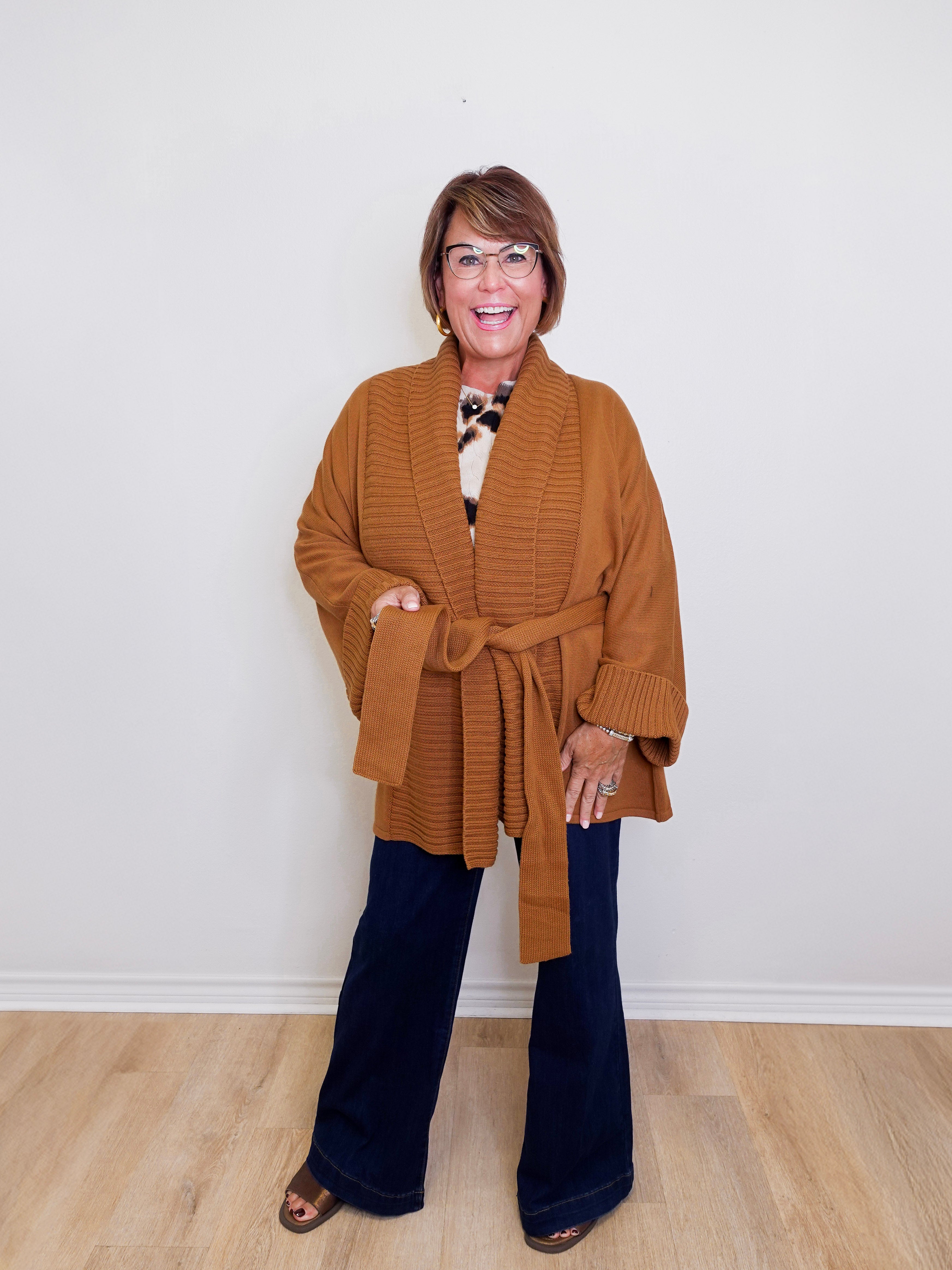 Woman wearing a brown cardigan, patterned top, and dark jeans against a white background