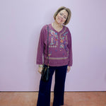 Woman wearing a purple embroidered top and dark pants against a light purple wall.