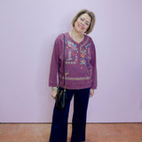 Woman wearing a purple embroidered top and dark pants against a light purple wall.
