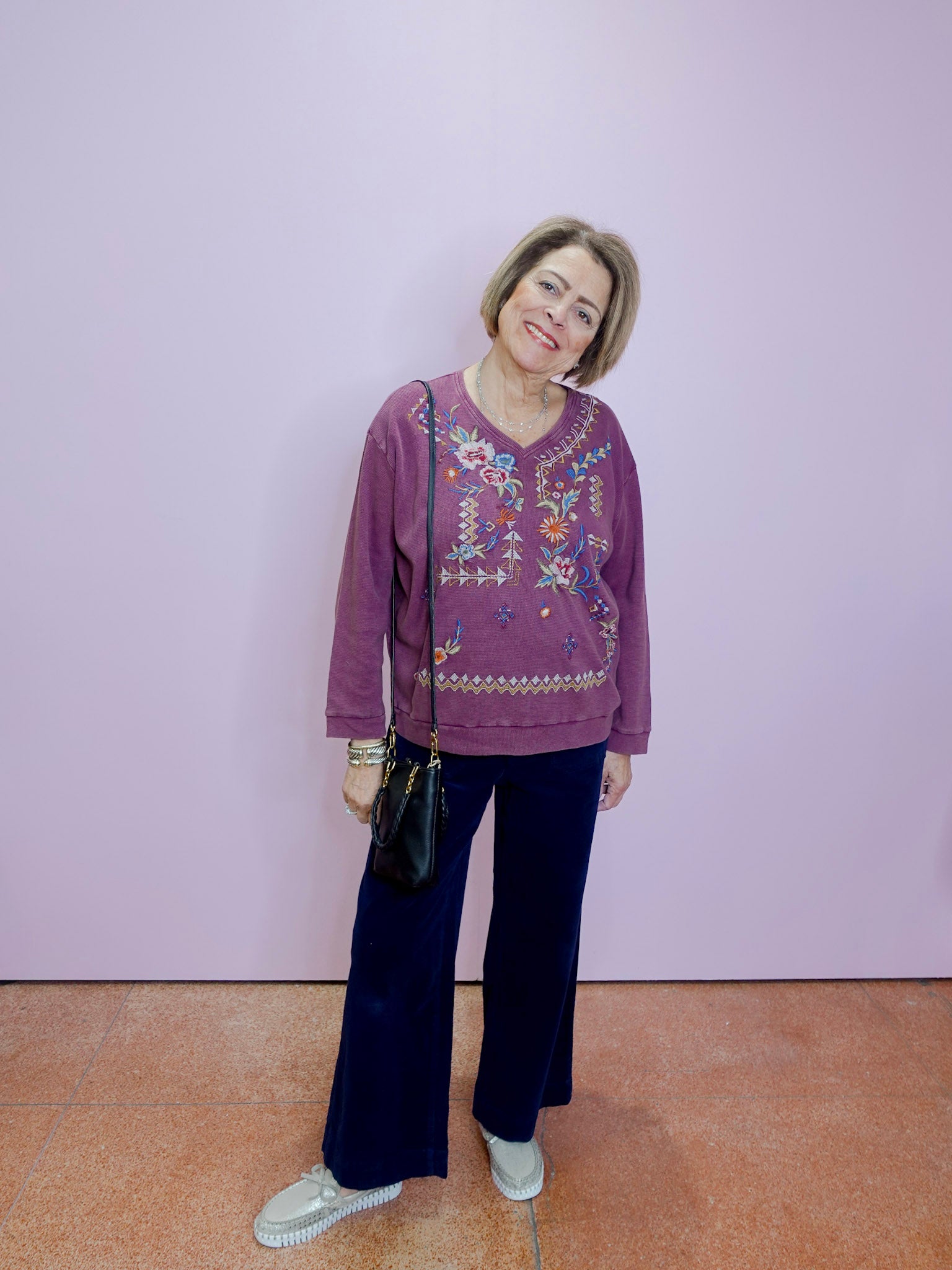 Woman wearing a purple embroidered top and dark pants against a light purple wall.
