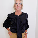 Woman wearing a black blouse with a bow tie and beige pants against a white background