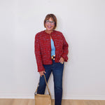 Woman wearing a red jacket, blue jeans, and holding a beige handbag against a white wall.