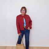Woman wearing a red jacket, blue jeans, and holding a beige handbag against a white wall.
