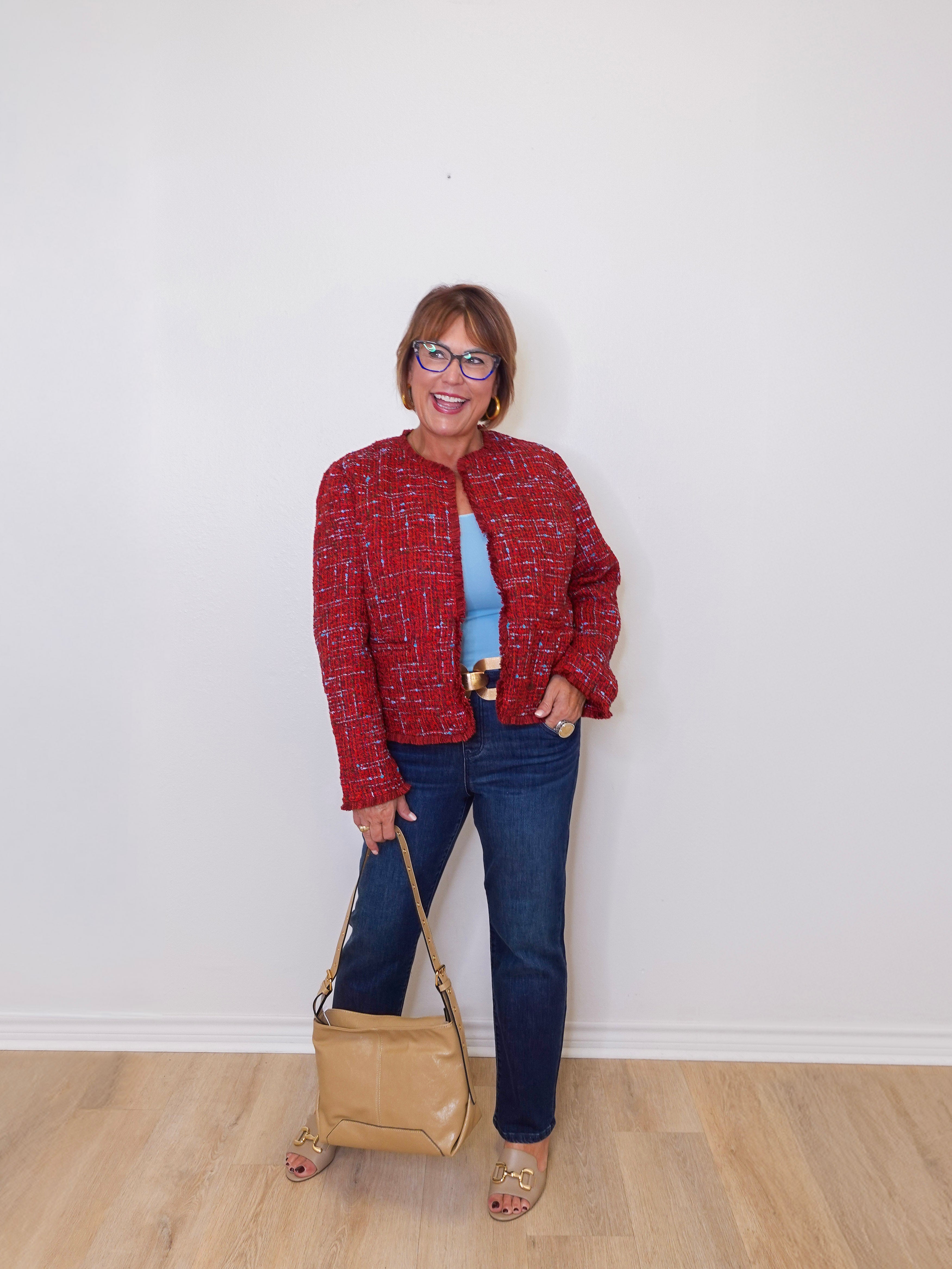Woman wearing a red jacket, blue jeans, and holding a beige handbag against a white wall.
