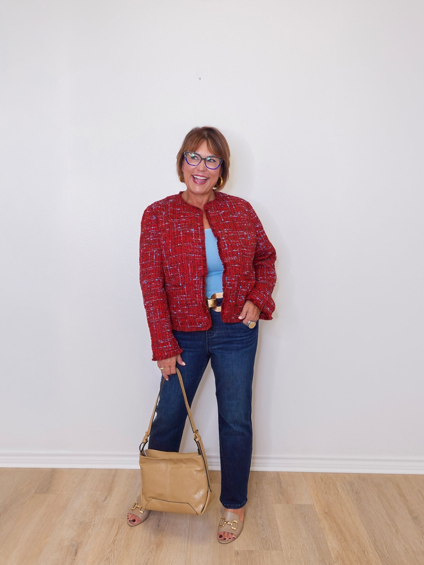 Woman wearing a red jacket, blue jeans, and holding a beige handbag against a white wall.