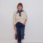 Woman wearing a beige sweater, blue jeans, and brown loafers against a white wall.