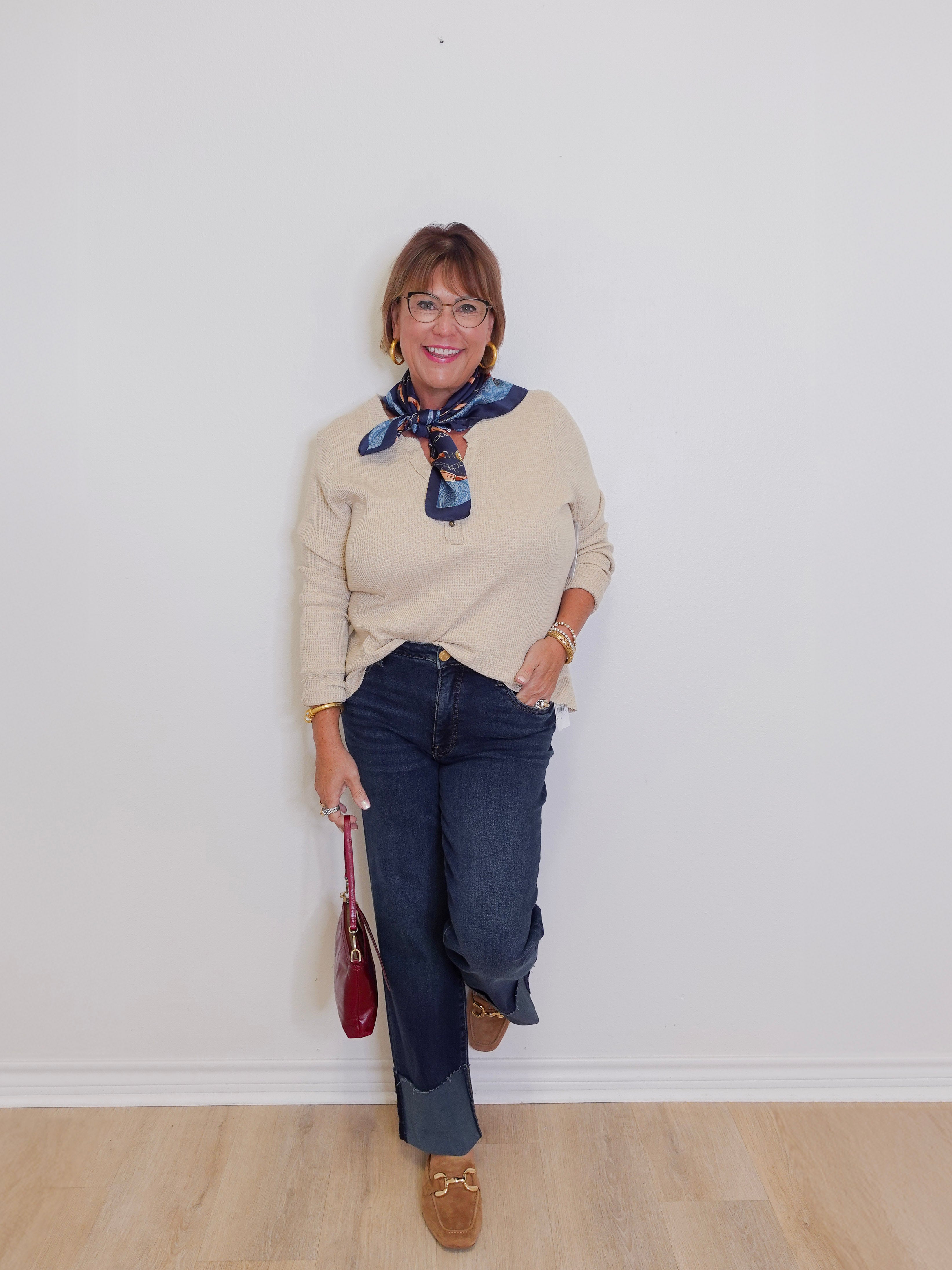 Woman wearing a beige sweater, blue jeans, and brown loafers against a white wall.