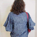Person wearing a blue patterned blouse with a red bag over one shoulder, standing against a white wall.