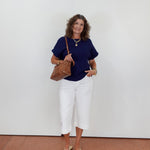 Woman wearing a navy top and white pants holding a brown bag against a white wall.