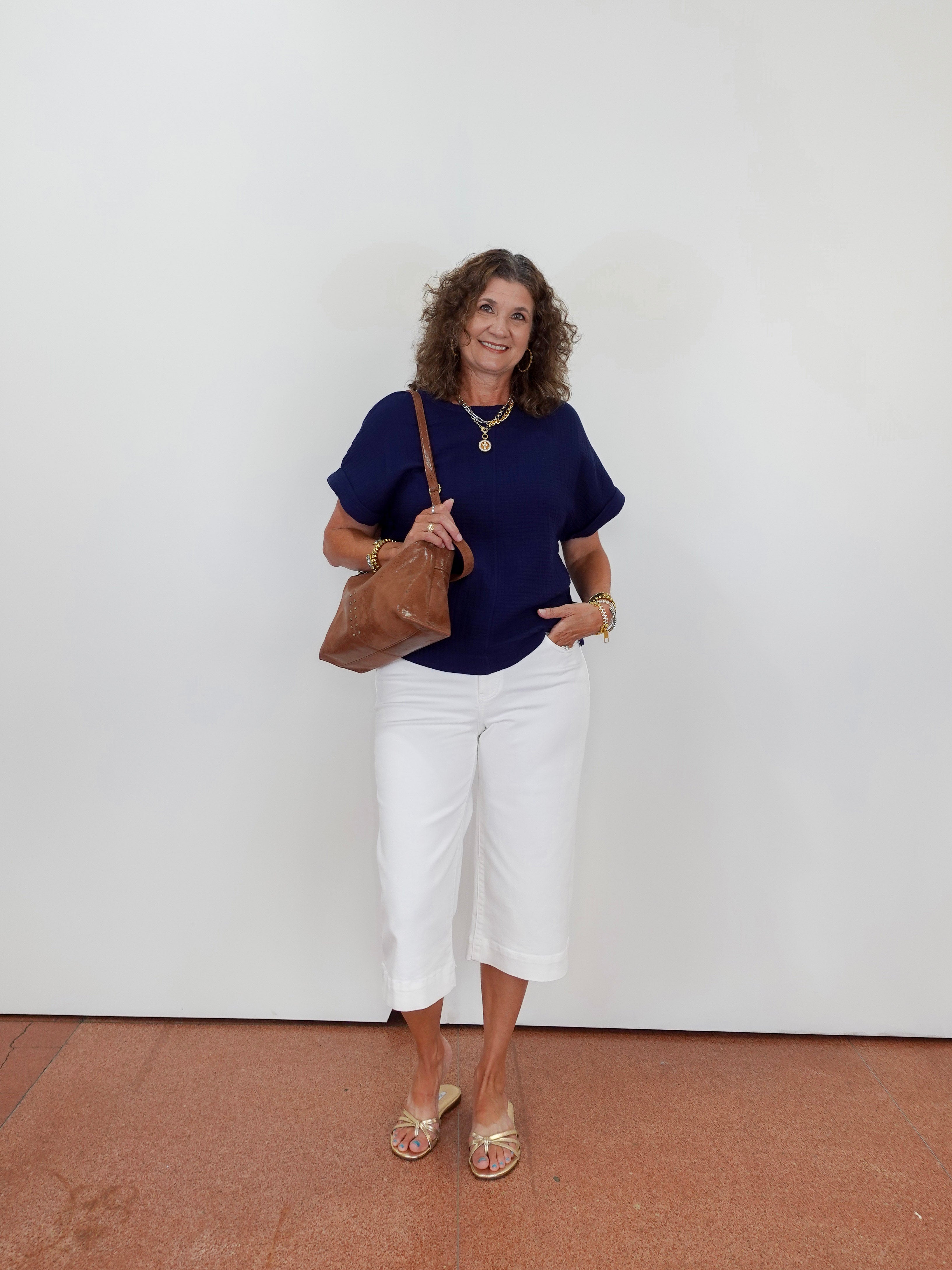 Woman wearing a navy top and white pants holding a brown bag against a white wall.