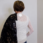 Person wearing a black floral jacket over a white lace top against a plain wall.