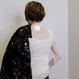 Person wearing a black floral jacket over a white lace top against a plain wall.