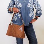 Person wearing a blue floral embroidered shirt and holding a brown handbag against a white background